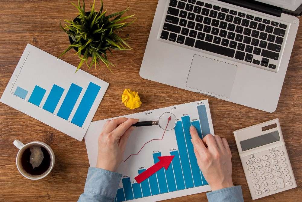 Top-down view of a person using a magnifying glass on a growth chart next to a laptop, calculator, and coffee cup.
