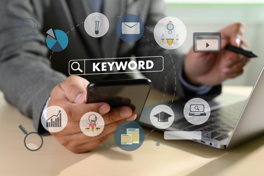 Keyword research concept with search bar and digital marketing icons above a smartphone and laptop workspace.